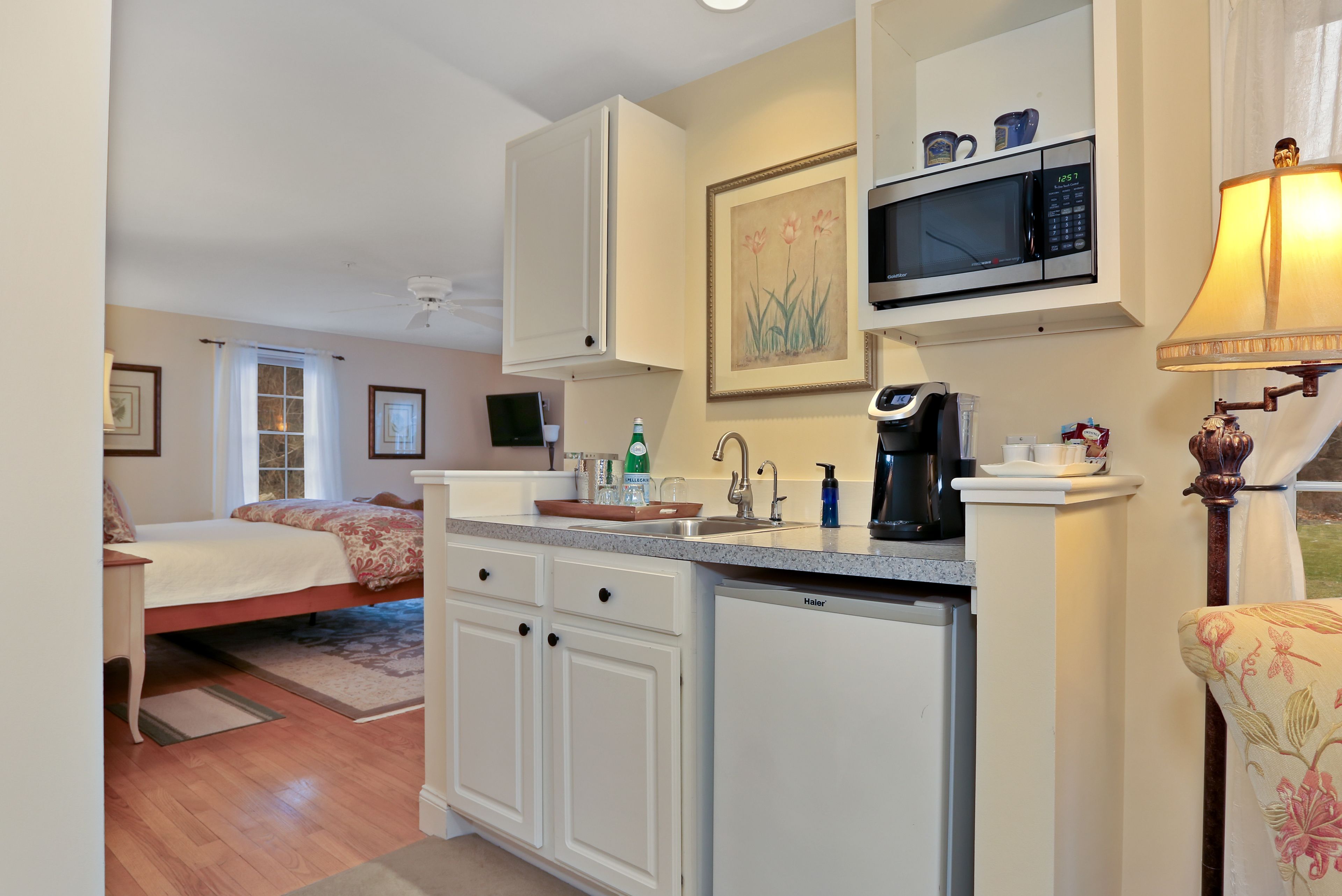 A compact bedroom with a bed, a small desk, and a nightstand. Adjacent to the bedroom is a kitchenette with a microwave, coffee maker, and a sink.