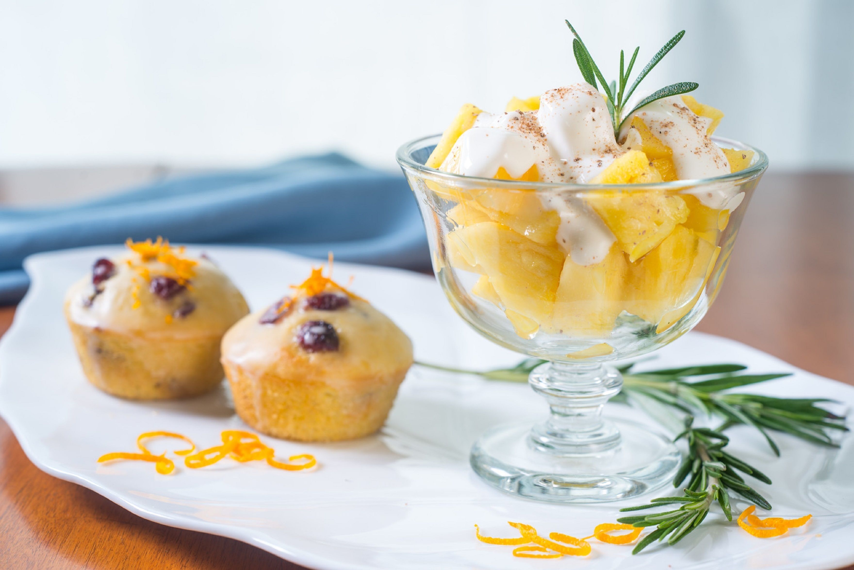 A dessert bowl with fruit and cream, accompanied by two muffins on a white plate, with a sprig of rosemary on the side.