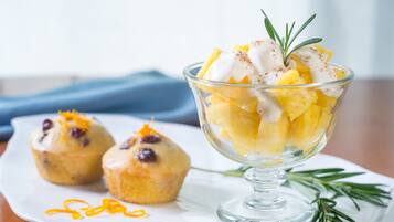 A dessert bowl with fruit and cream, accompanied by two muffins on a white plate, with a sprig of rosemary on the side.