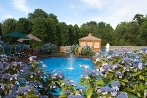 En swimmingpool med en fontän, omgiven av frodig grönska och en lusthus.