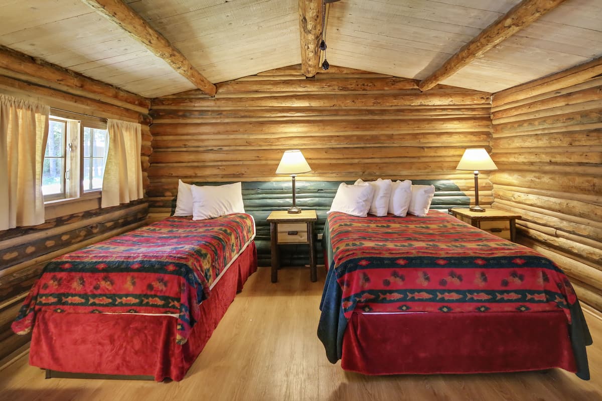 A log cabin room with two beds, each with a patterned bedspread, a small table, and a lamp between the beds.