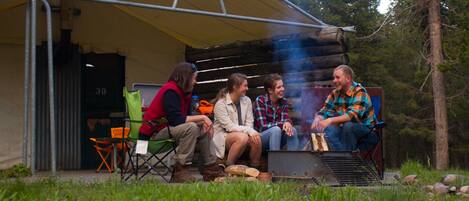 A group of people sitting outside a covered structure with a grill, surrounded by greenery.