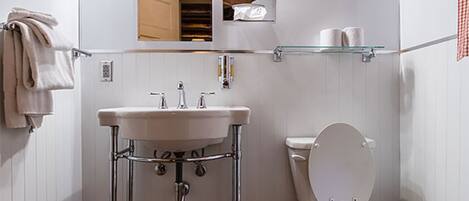 A bathroom with a wooden ceiling, white walls, a white sink, a toilet, and a glass shelf above the toilet.
