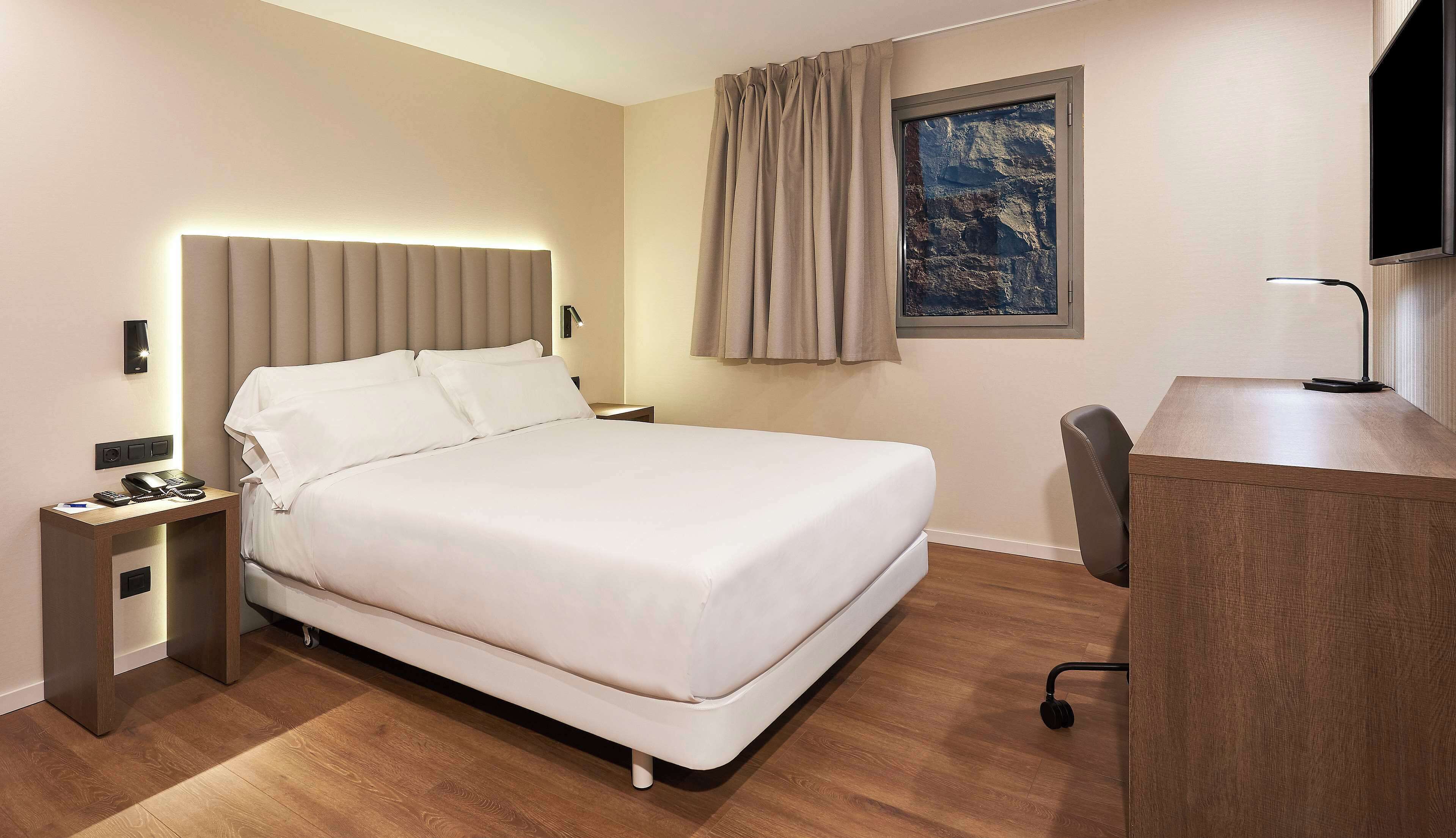 Une chambre d’hôtel avec un lit, une table de chevet, une lampe, un bureau, une chaise et un tableau au mur.