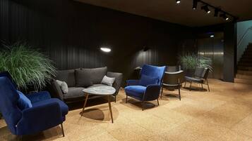 A modern lounge area with a dark-colored sofa, blue armchairs, and a small round table. There are potted plants and a staircase in the background.