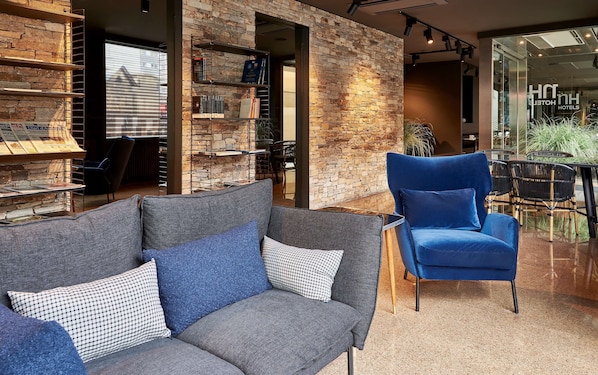 A modern hotel lobby with a stone wall, a blue armchair, a grey sofa, and a glass door leading outside. - NH Andorra la Vella (Andorra la Vella)