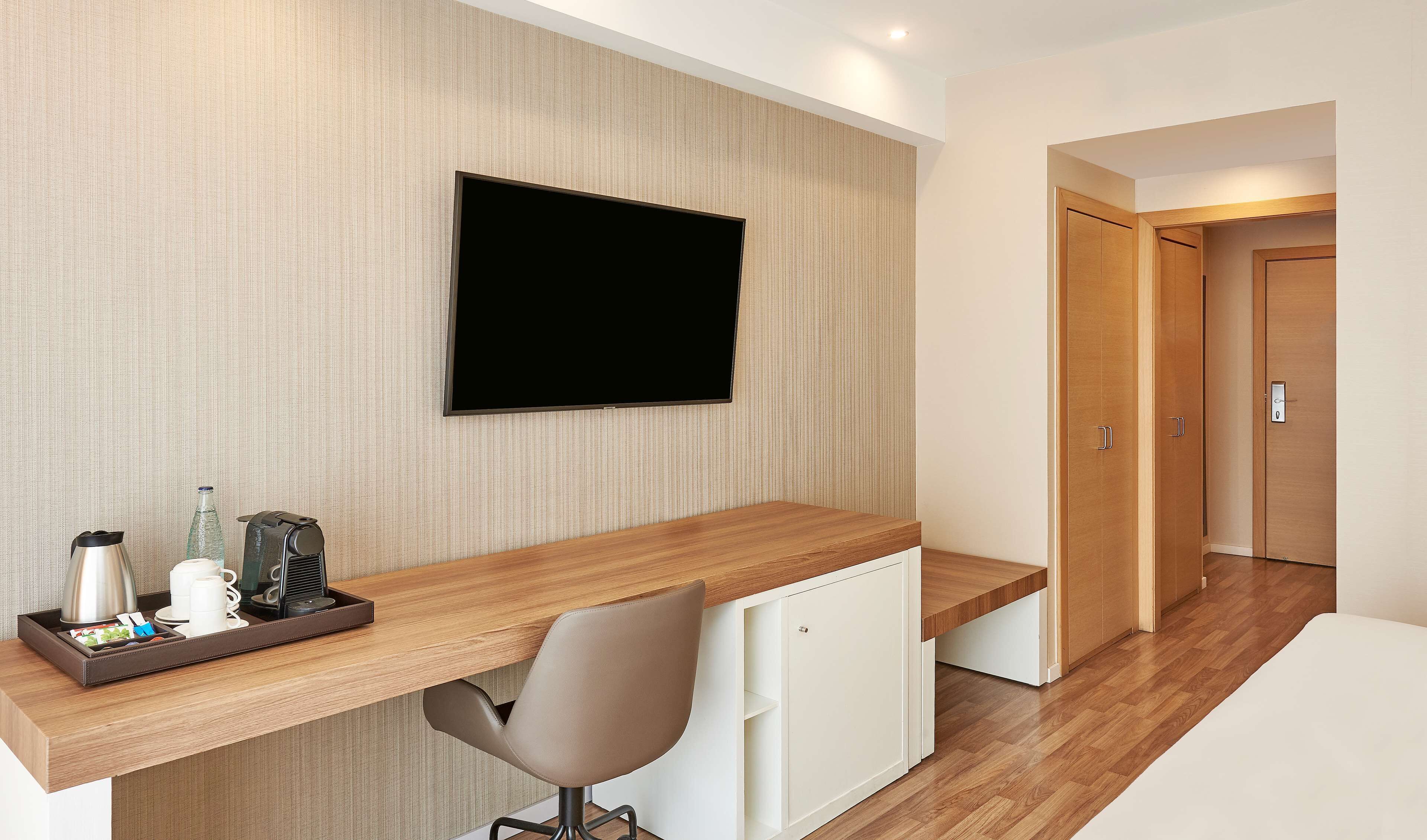 A modern hotel room with a wooden desk, a flat-screen TV, a chair, and a small kitchenette.