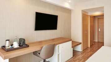 A modern hotel room with a wooden desk, a flat-screen TV, a chair, and a small kitchenette.