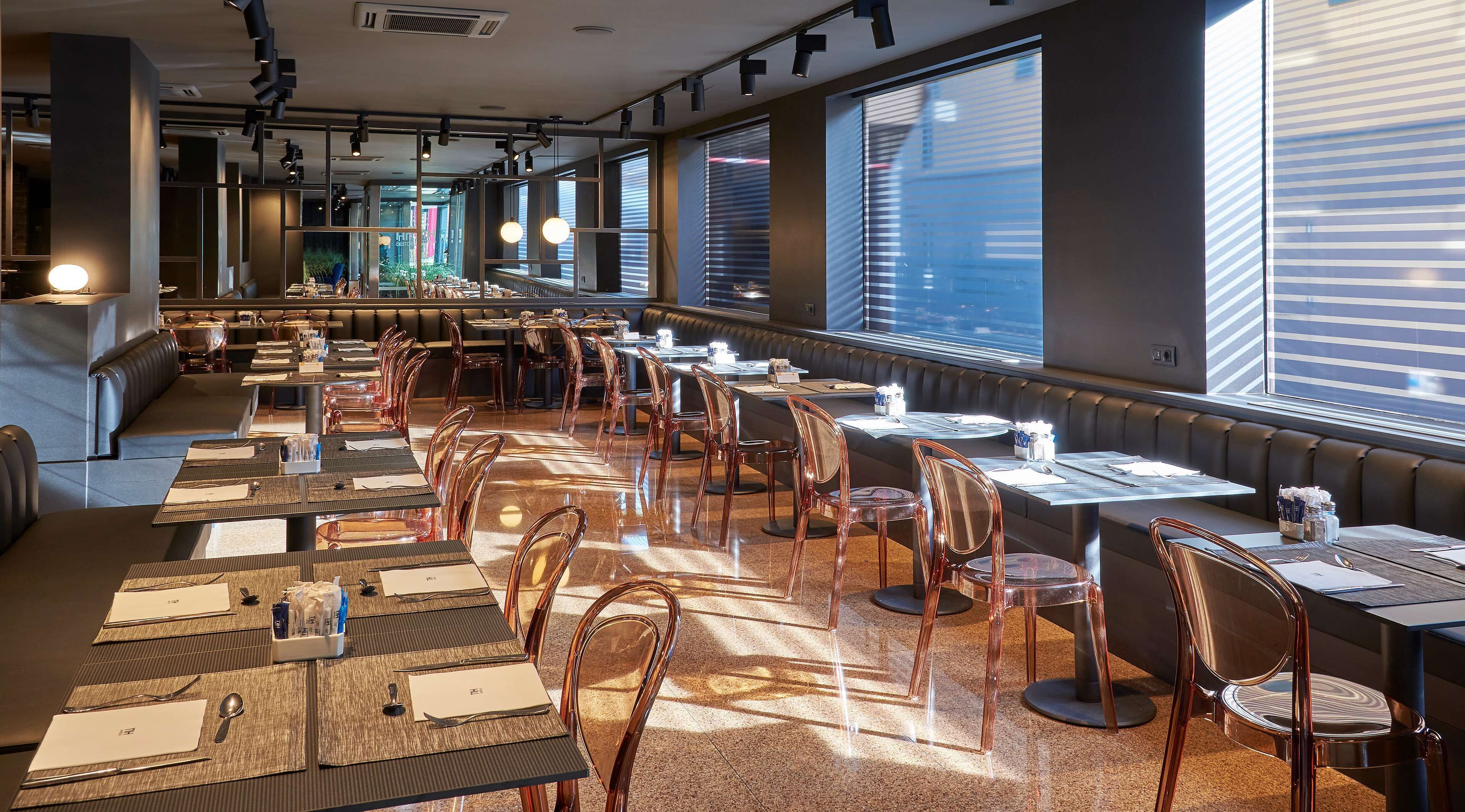 A restaurant with long communal tables, clear chairs, and a view of the city through large windows.