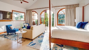 Quarto espaçoso com uma cama grande, um sofá e uma mesinha. Janelas amplas com cortinas, um ventilador de teto e vista para o verde lá fora.