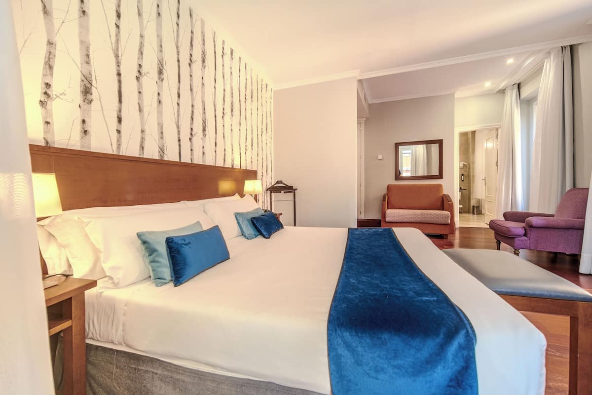 A hotel room with a large bed, a wooden headboard, a nightstand with a lamp, a seating area with a chair, and a bathroom visible through an open door.