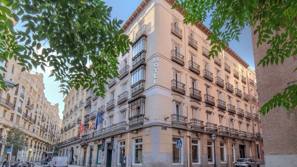 Um hotel com vários andares, varandas, placa indicando ser um hotel e uma área de pedestres com veículos estacionados.