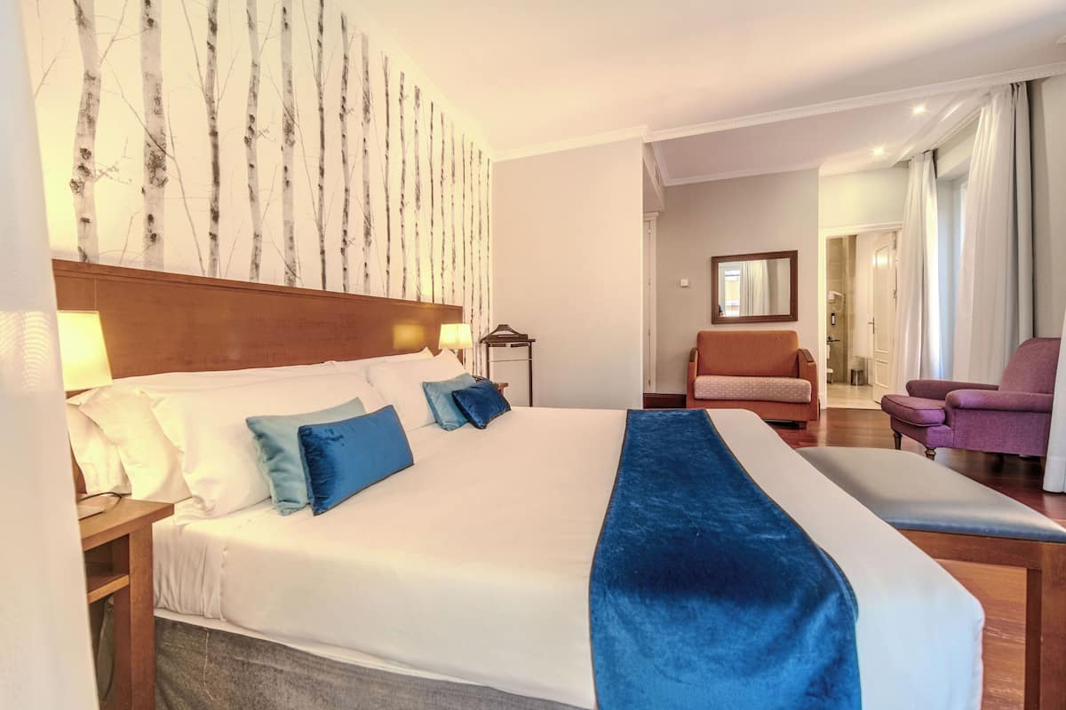 A hotel room with a large bed, a wooden headboard, a nightstand, a desk, a chair, and a bathroom visible through an open door.