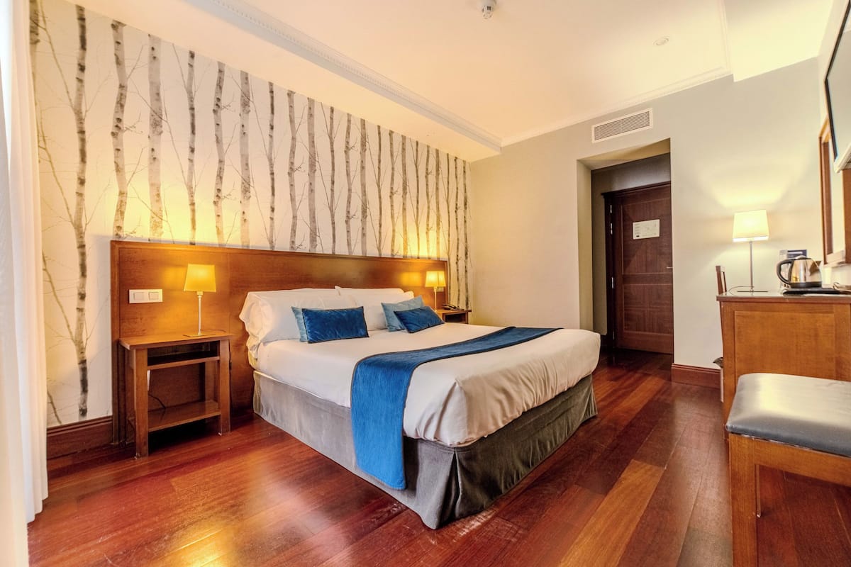 A hotel room with a large bed, wooden headboard, two bedside tables with lamps, a wooden desk, and a chair.