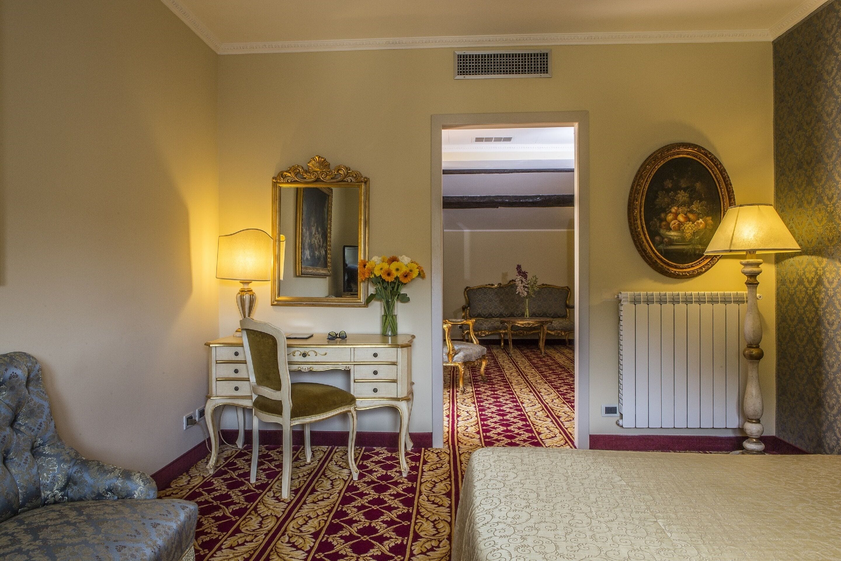 A hotel room with a bed, a desk with a chair, a dresser, a mirror, a radiator, and a lamp.