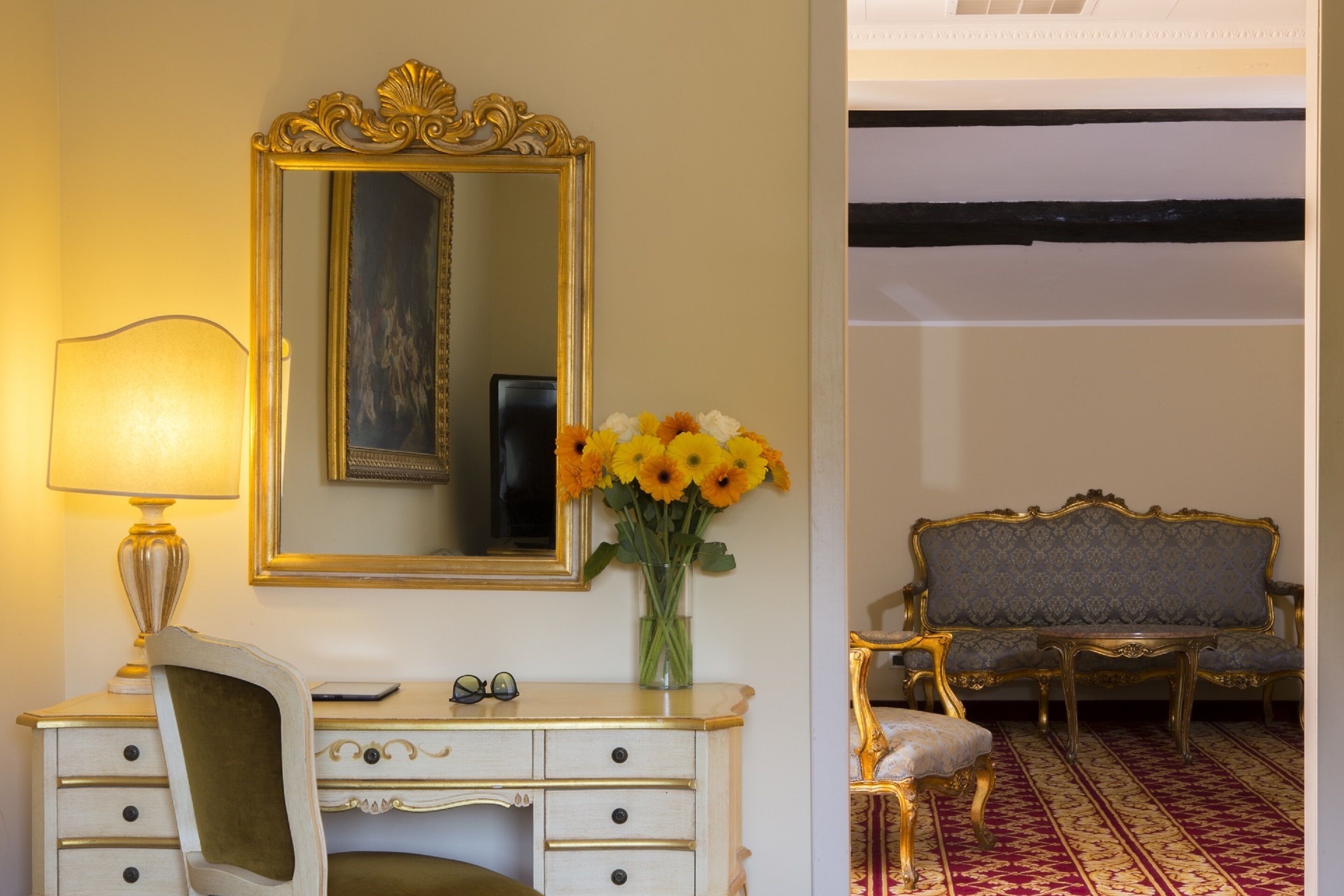 Una habitación con un tocador blanco, un espejo con marco dorado, un florero con flores amarillas, una lámpara, un par de gafas y un sofá tapizado azul con un alfombra estampado.