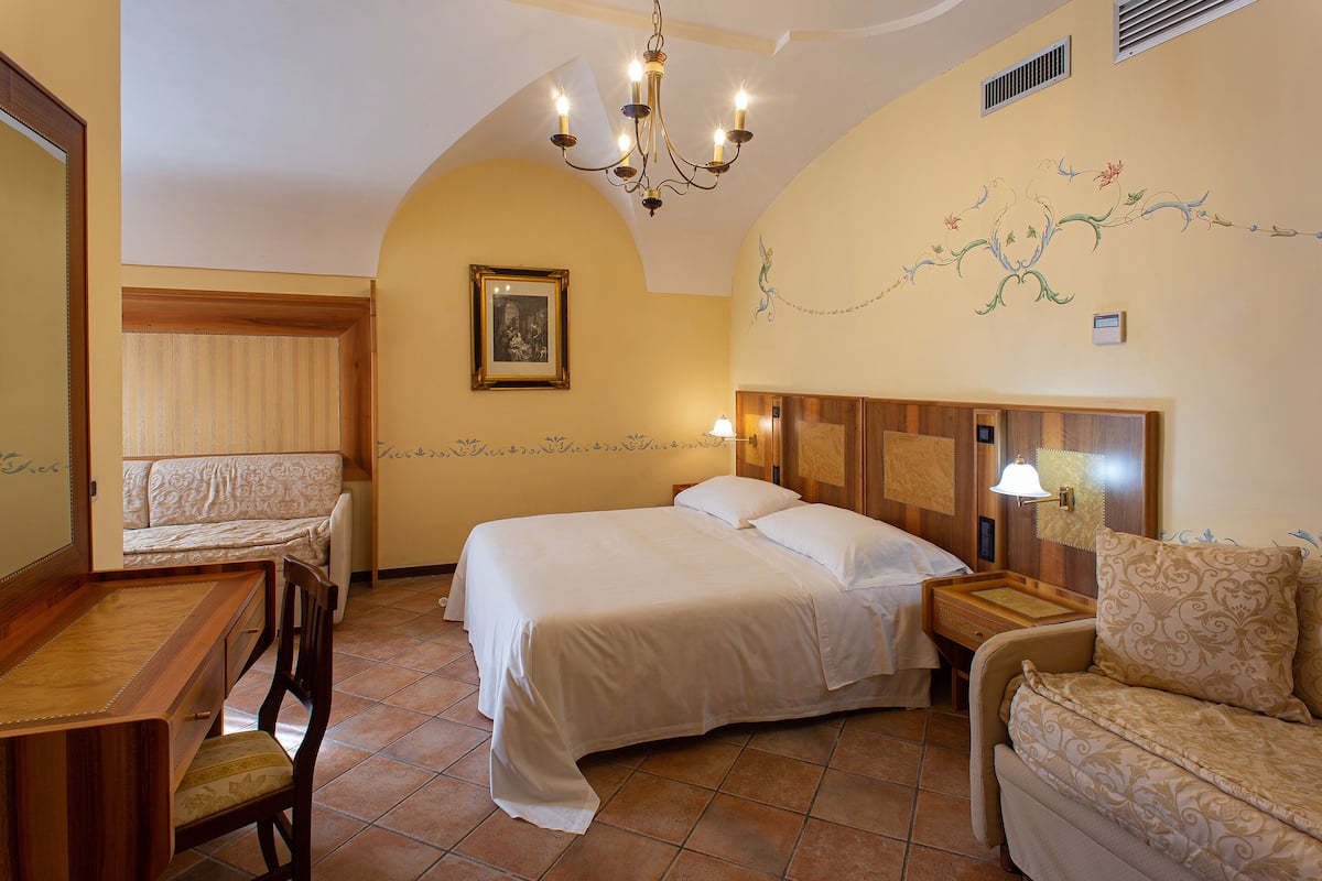 Hotelkamer met een bed, bureau, stoel, bank, een kroonluchter en een wand met bloemenmotief.