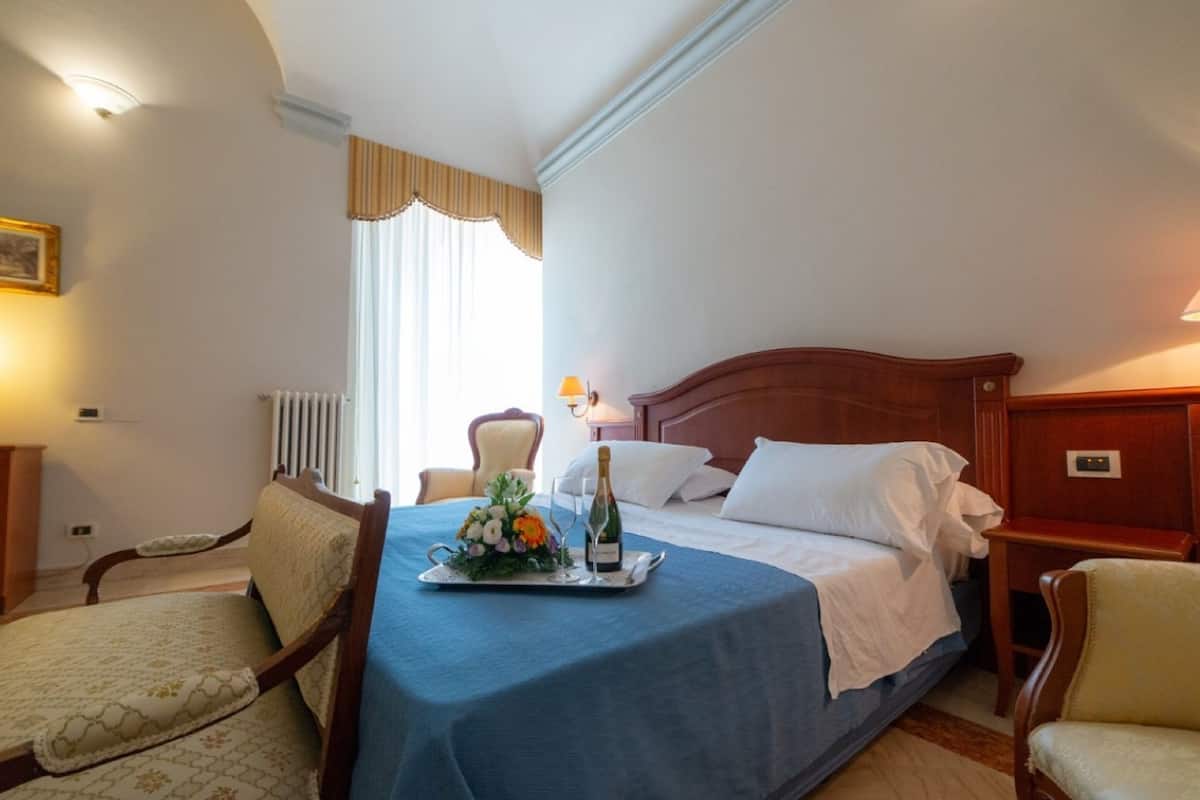 Hotelkamer met een bed, een stoel, een tafel met een schaal bloemen en een fles, en een raam met gordijnen.