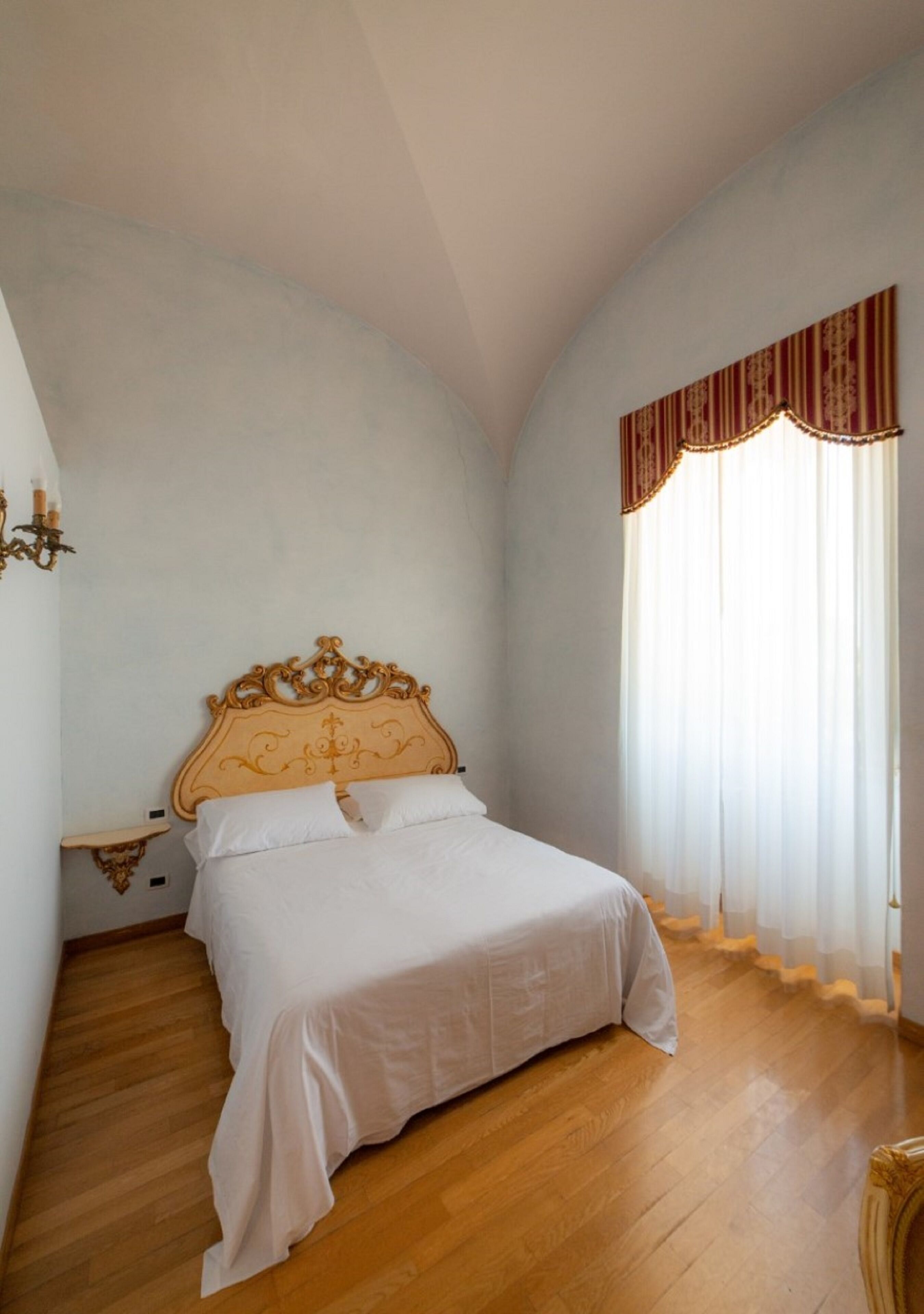 Un dormitorio con una cama de madera, ropa de cama blanca, una mesita de noche y una ventana con cortinas traslúcidas.