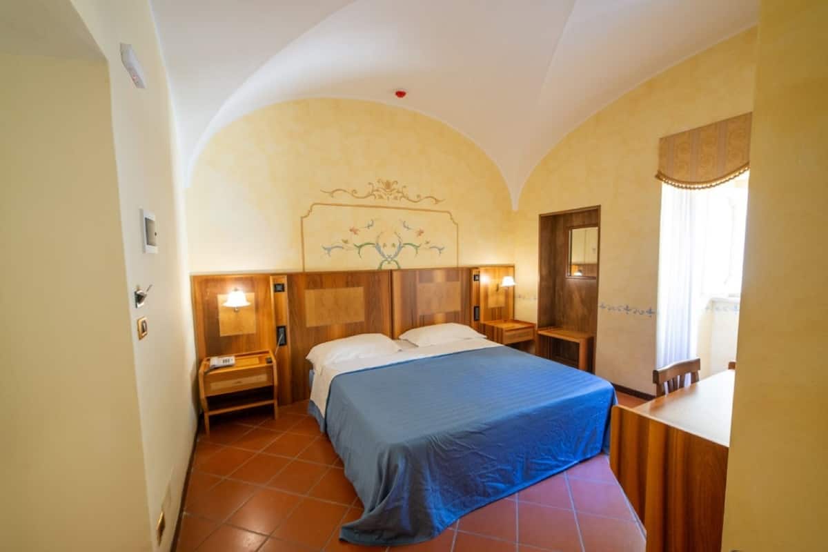 Een hotelkamer met een bed, nachtkastje, spiegel en raam met gordijnen.
