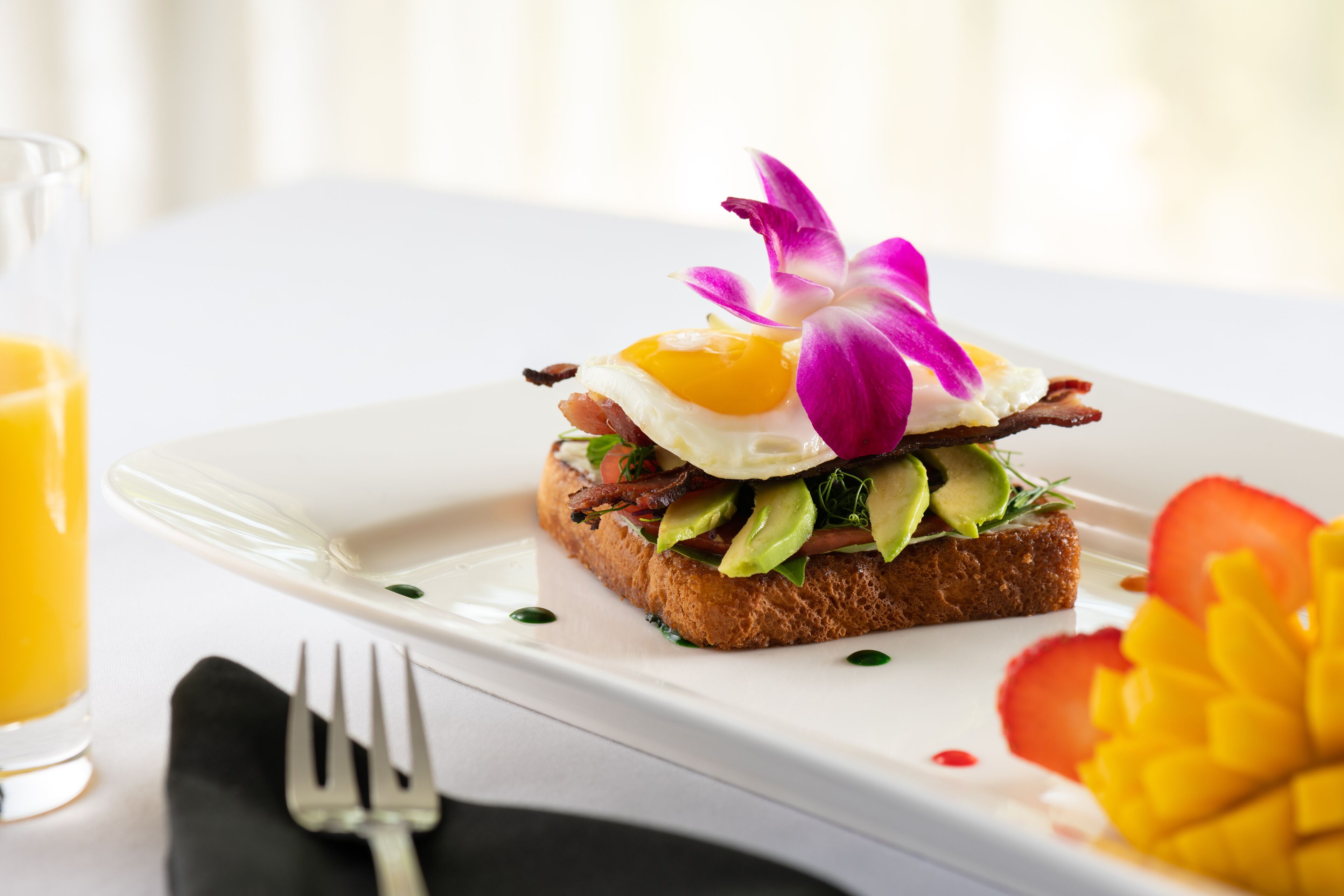 A breakfast plate with toast, egg, avocado, and bacon, garnished with a purple flower, on a white plate.
