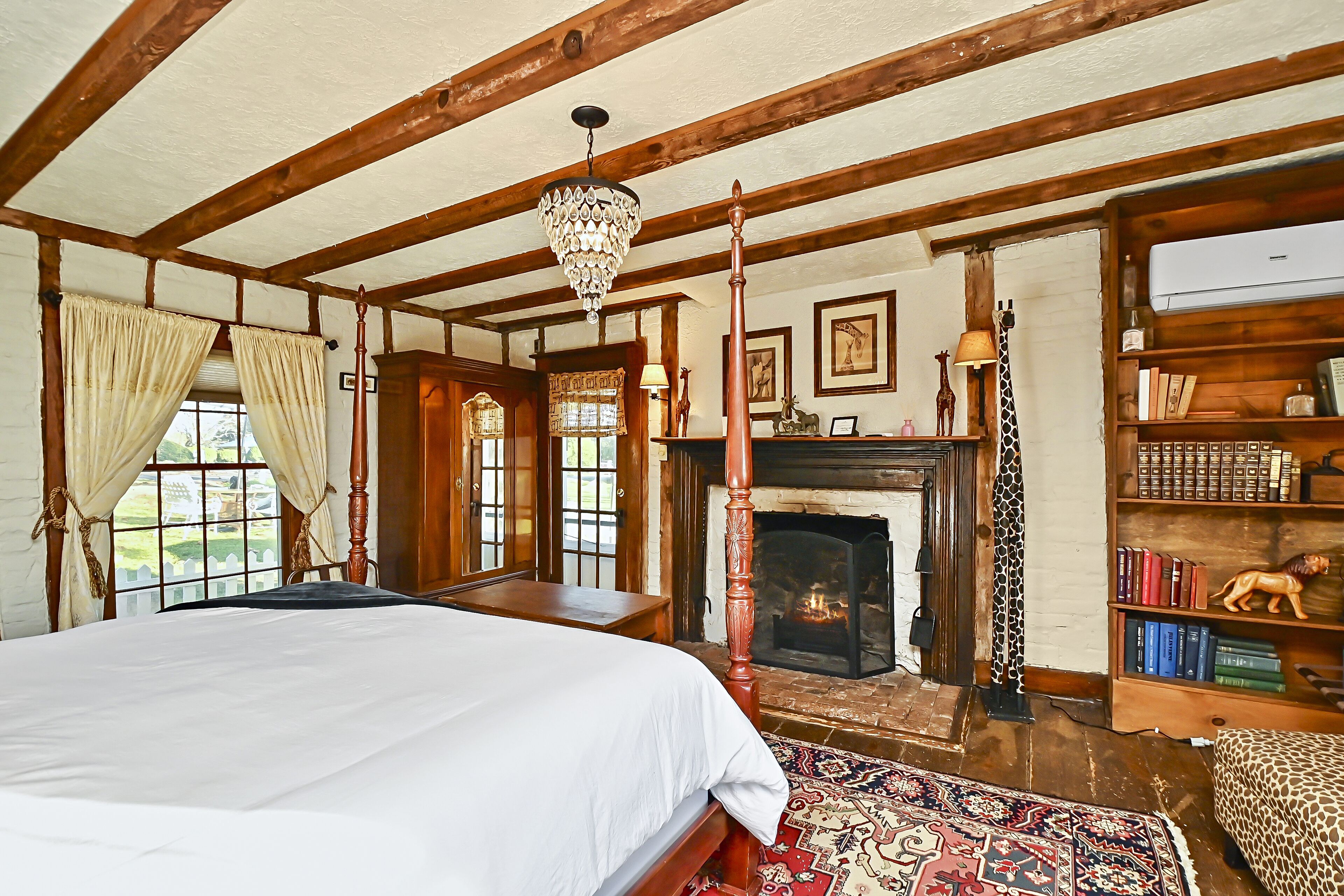 A bedroom with a large bed, wooden beams, a fireplace, a bookshelf, and a view of the outside.