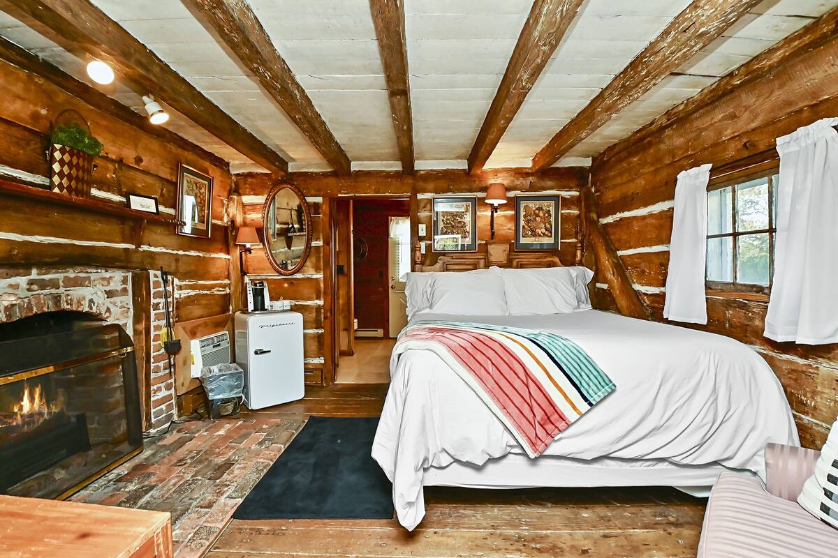 Un dormitorio rústico de cabaña de madera con chimenea, una cama con una manta colorida y un refrigerador pequeño.