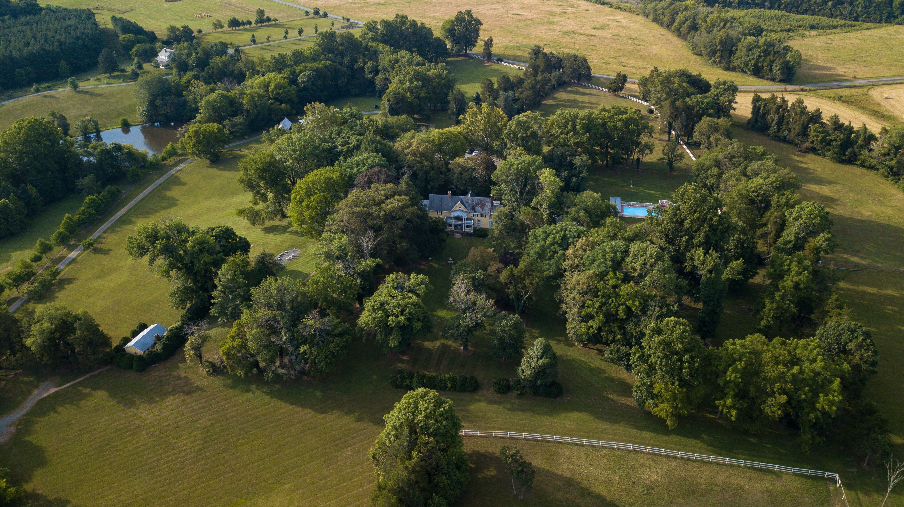 An aerial view of a large estate with a main house surrounded by dense trees, a swimming pool, and a fenced area.