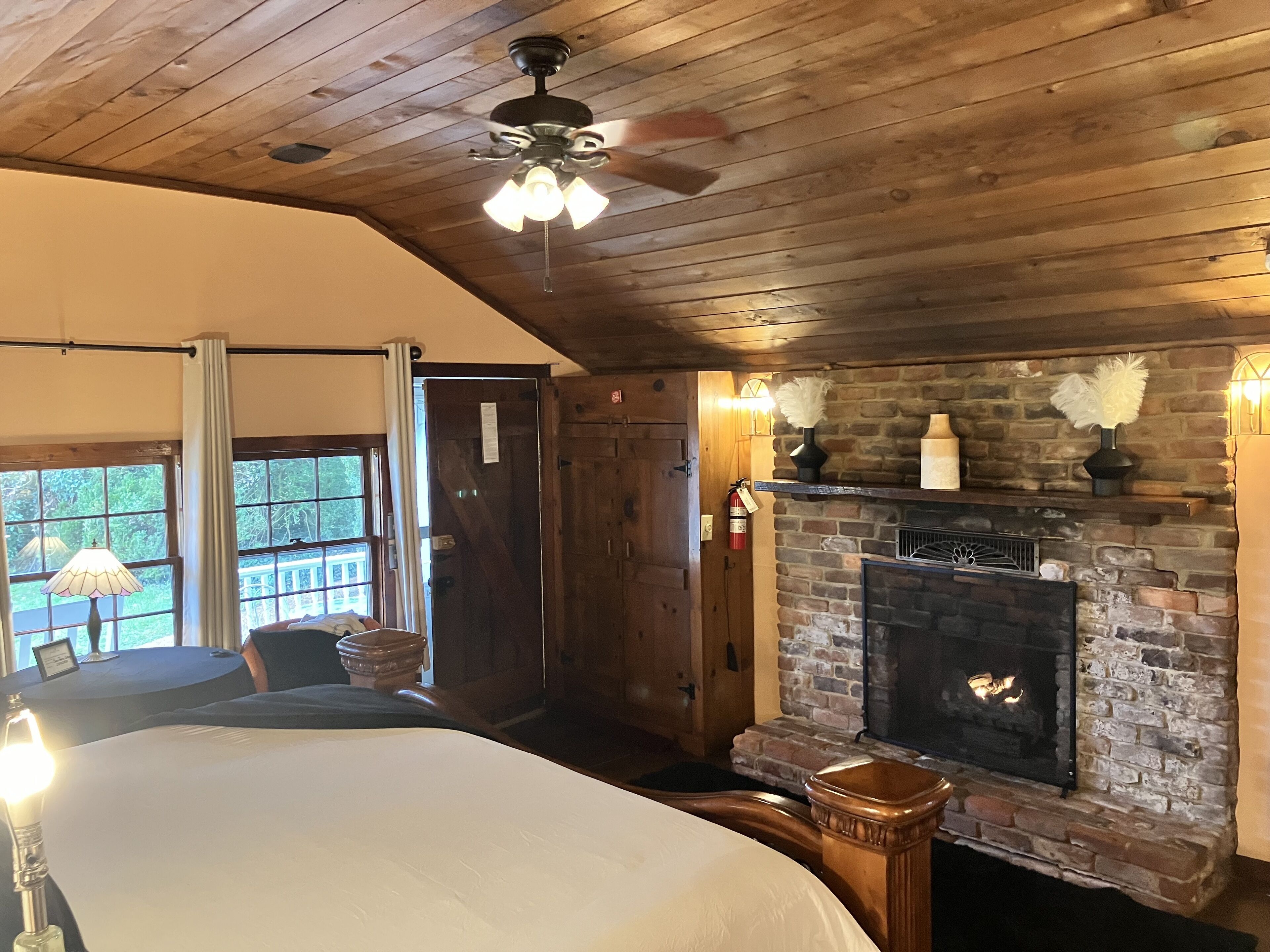A cozy room with a wooden ceiling, a fireplace, a bed, and a table with a lamp.
