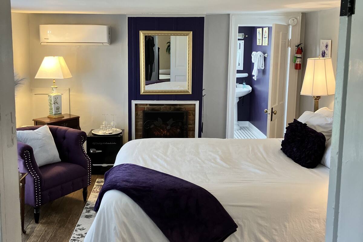 Una habitación de hotel con una cama grande, una silla, una mesita de noche, un espejo, una chimenea y un baño visible a través de una puerta abierta.