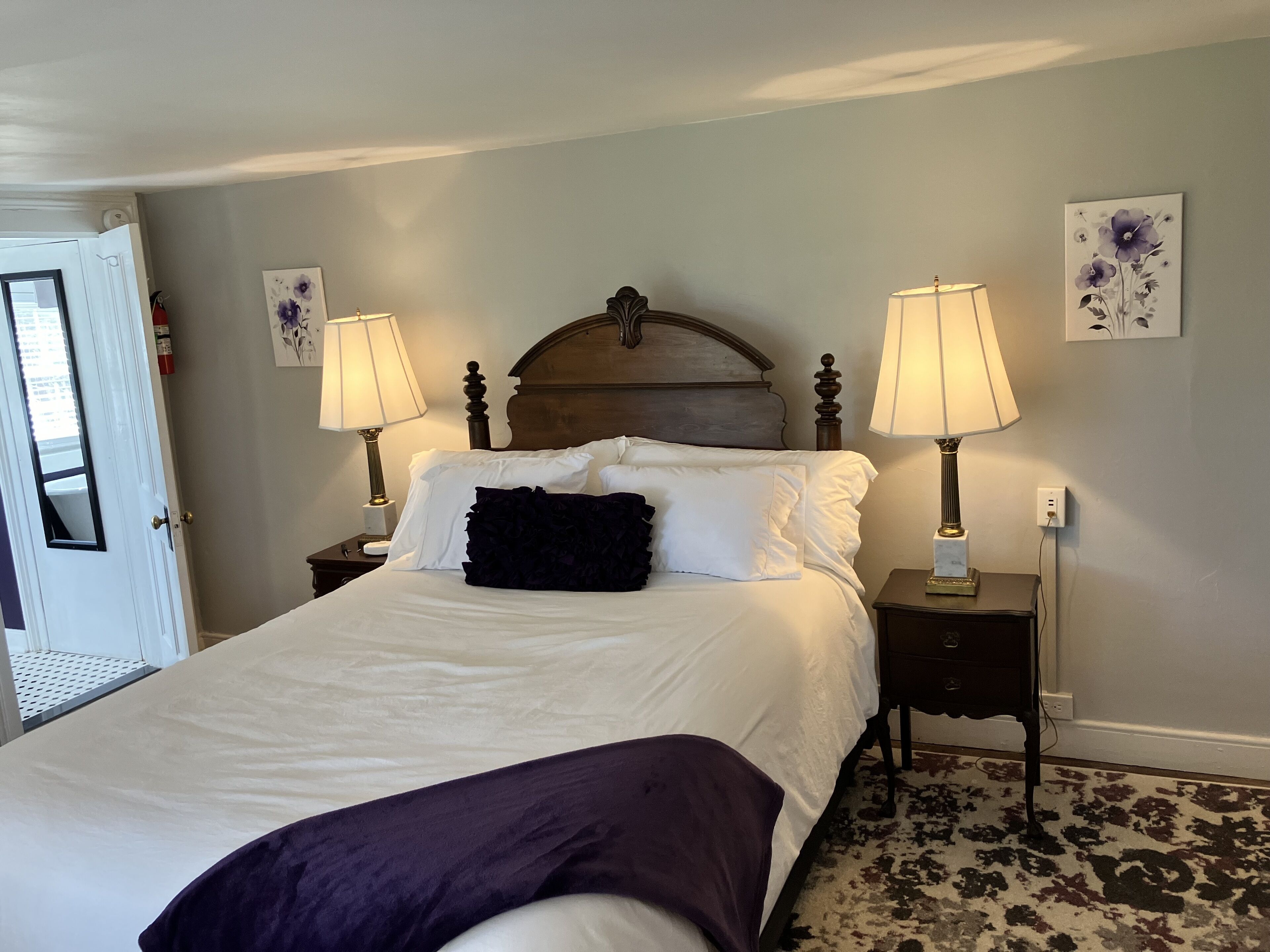 A neatly made bed with white linens and a dark throw blanket, flanked by two bedside tables with lamps, in a room with a patterned carpet and light-colored walls.