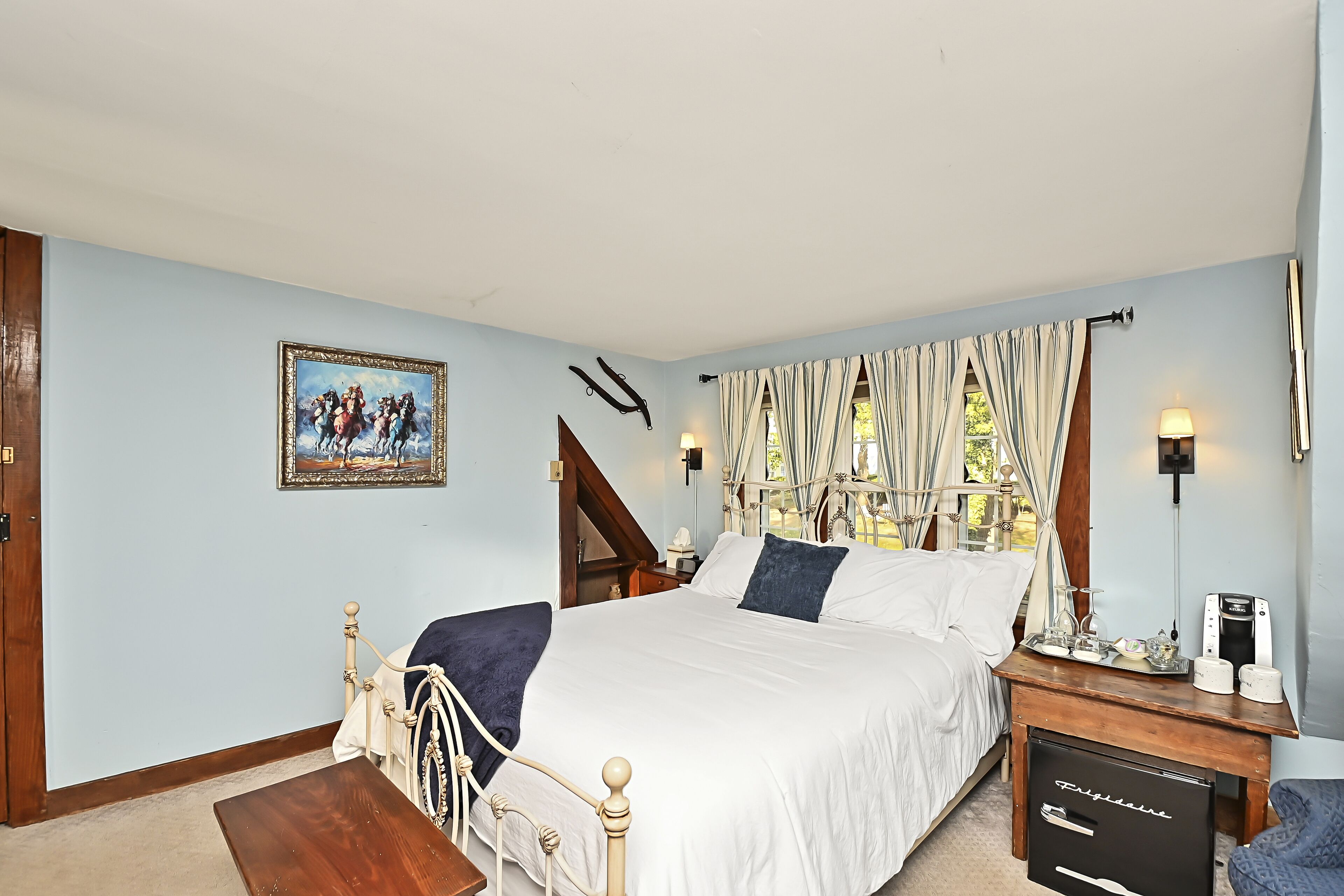 Une chambre à coucher avec un grand lit, une table de chevet en bois, un bureau avec une lampe, un tableau au mur et une fenêtre avec des rideaux.