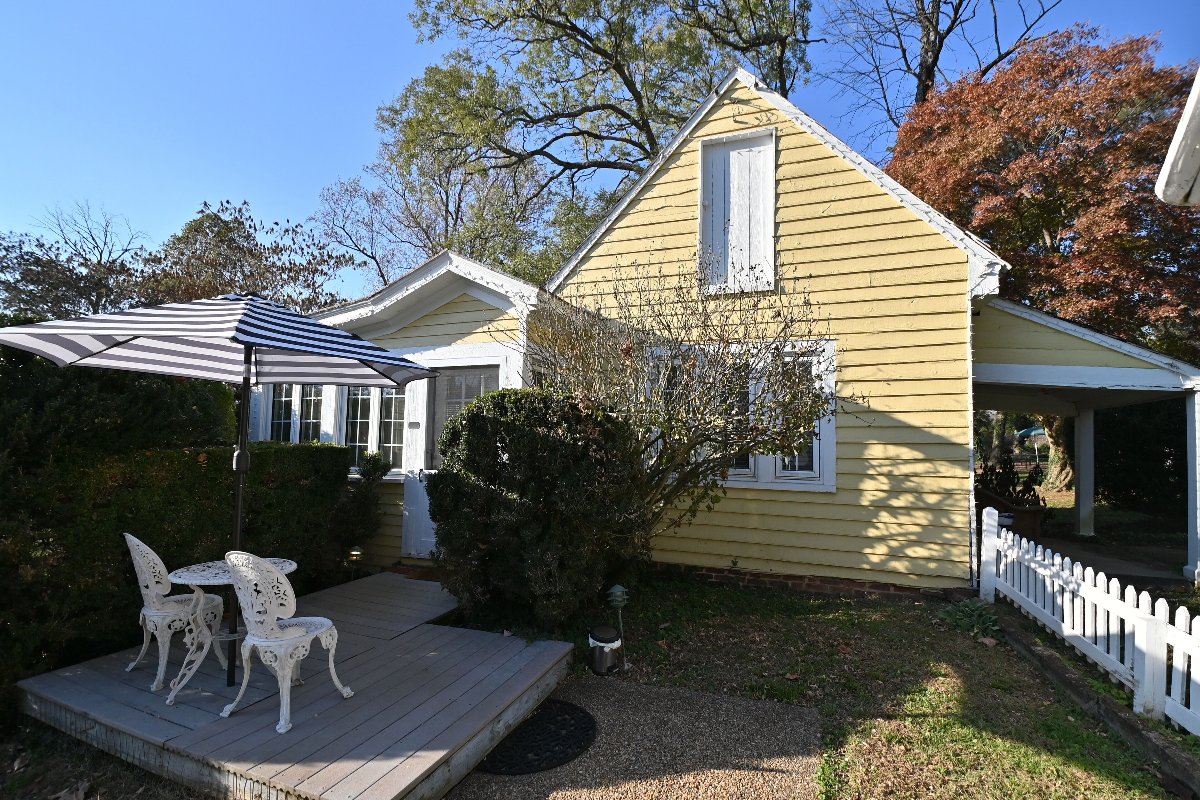 A yellow house with a porch, a small deck with white chairs, and a white picket fence.