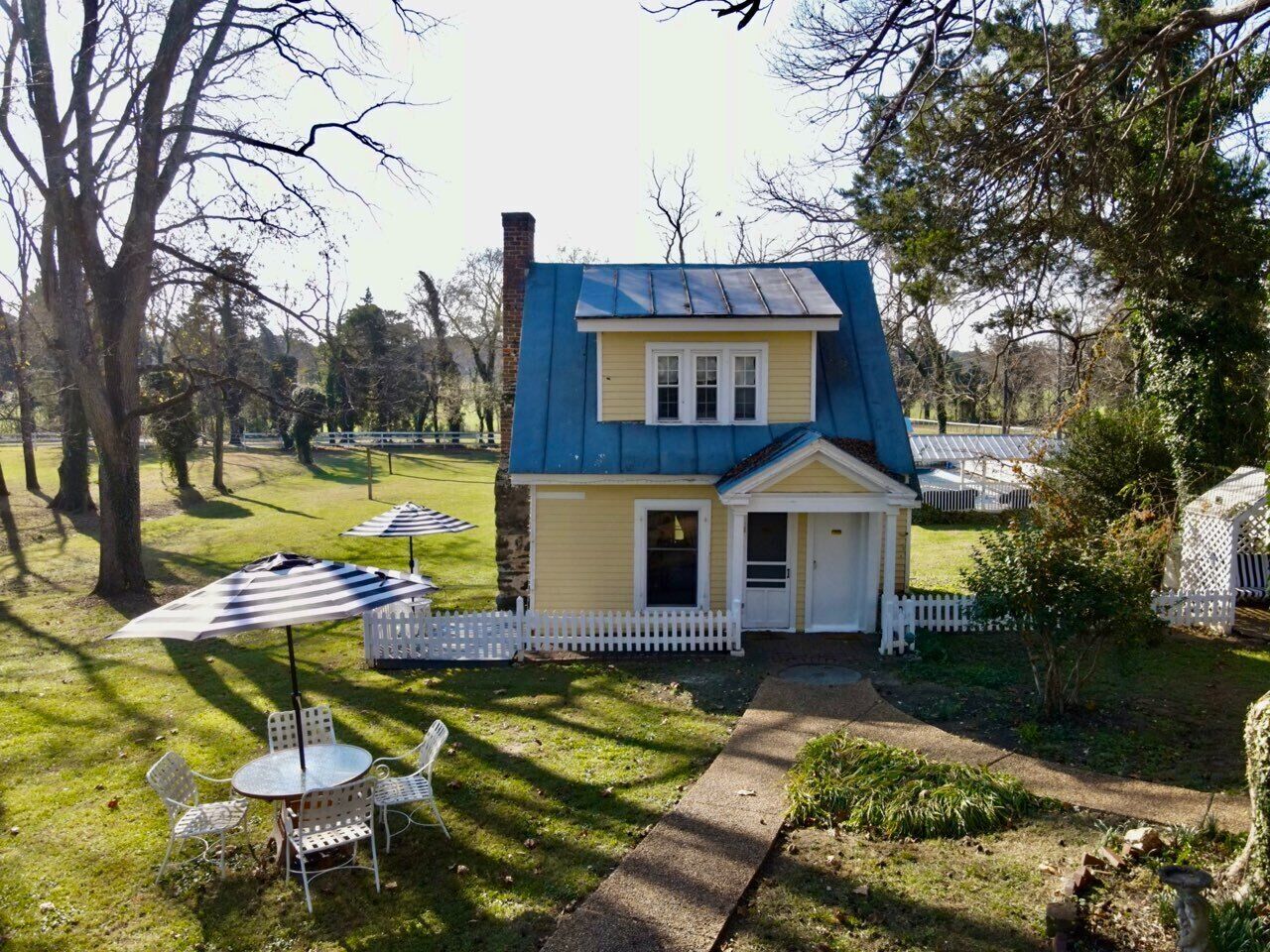 A small, single-story house with a blue roof and a white picket fence. There are outdoor tables and chairs, and a pathway leading to the house.