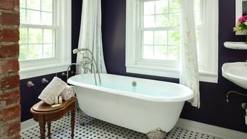 A bathroom with a freestanding bathtub, a wooden stool with toiletries, and a window with a view of greenery.