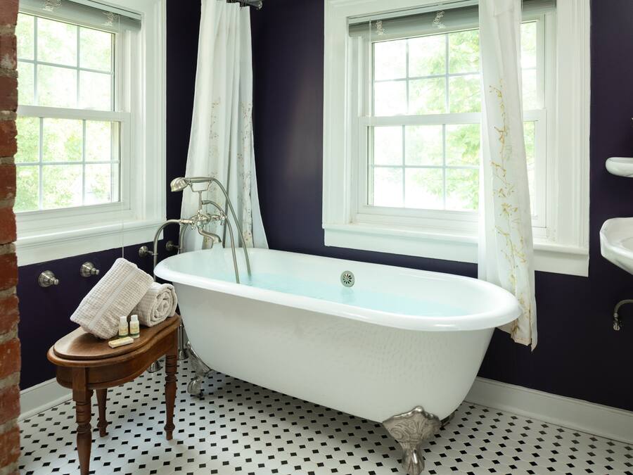 A bathroom with a freestanding bathtub, a wooden stool with toiletries, and a window with a view of greenery.