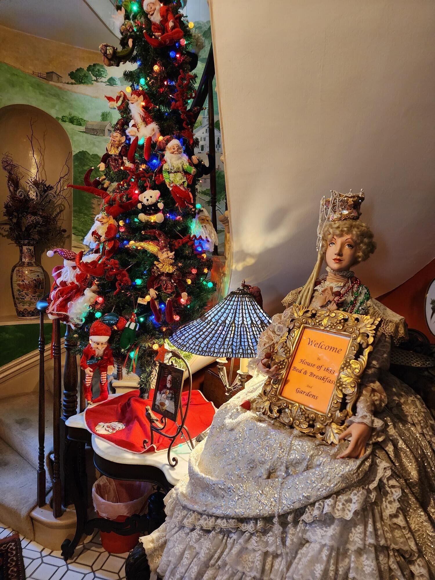 A Christmas tree with various ornaments and a doll dressed in a historical gown holding a sign.