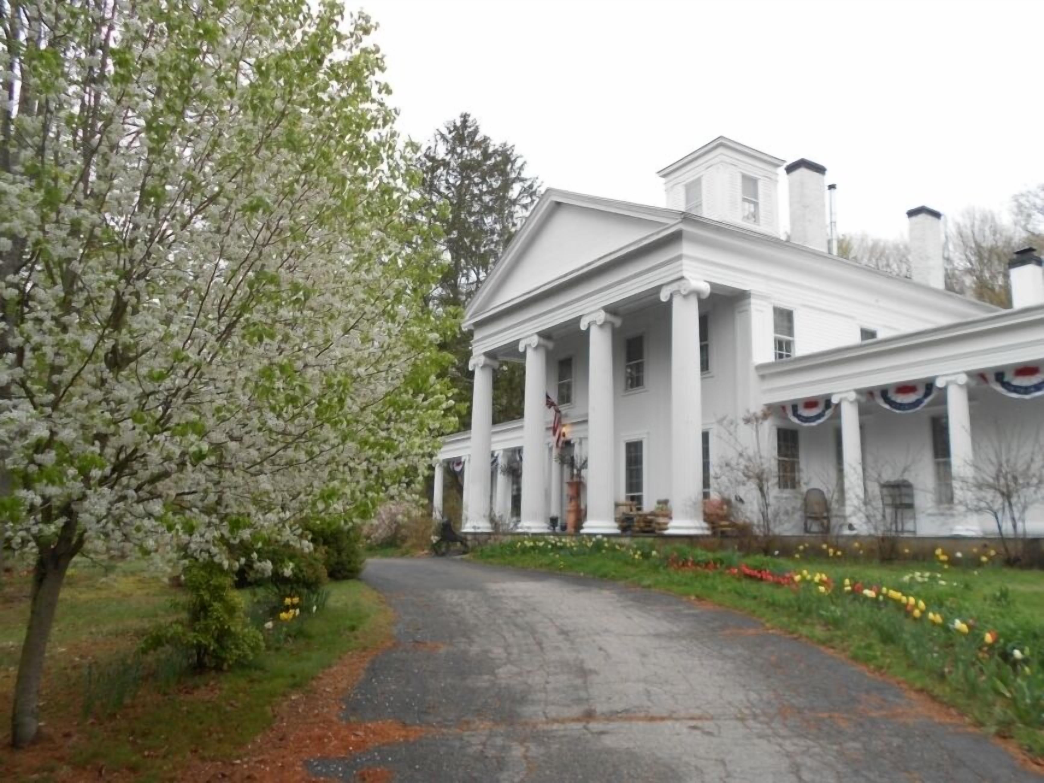 A white colonial-style building with a central portico, surrounded by a garden with blooming trees and flowers.