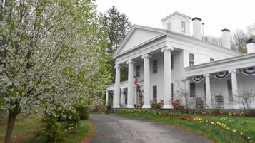 A white colonial-style building with a central portico, surrounded by a garden with blooming trees and flowers.
