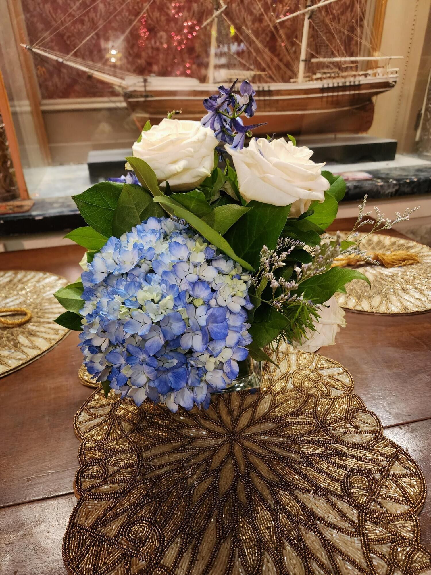 Eine dekorative Vase mit perlgarnierter Basis, gefüllt mit weißen Rosen, blauen Hortensien und violetten Blüten, auf einer gemusterten Matte platziert.