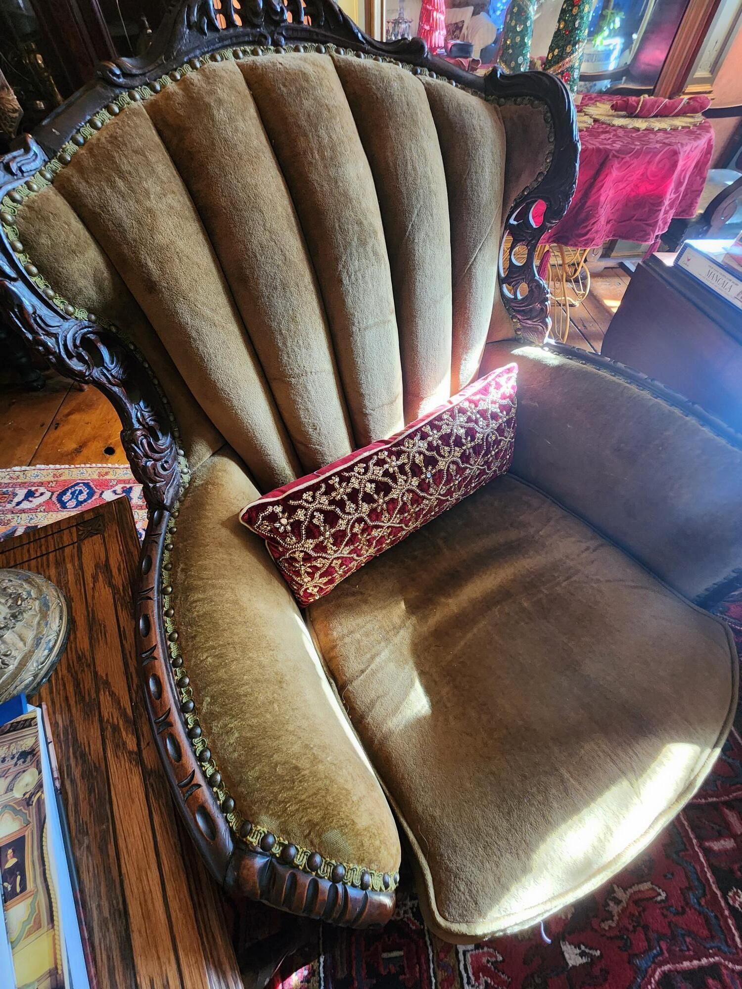 Sillón de estilo antiguo con respaldo alto y elaborada estructura de madera, con un asiento tapizado de color marrón claro y un cojín decorativo rojo con bordados dorados.