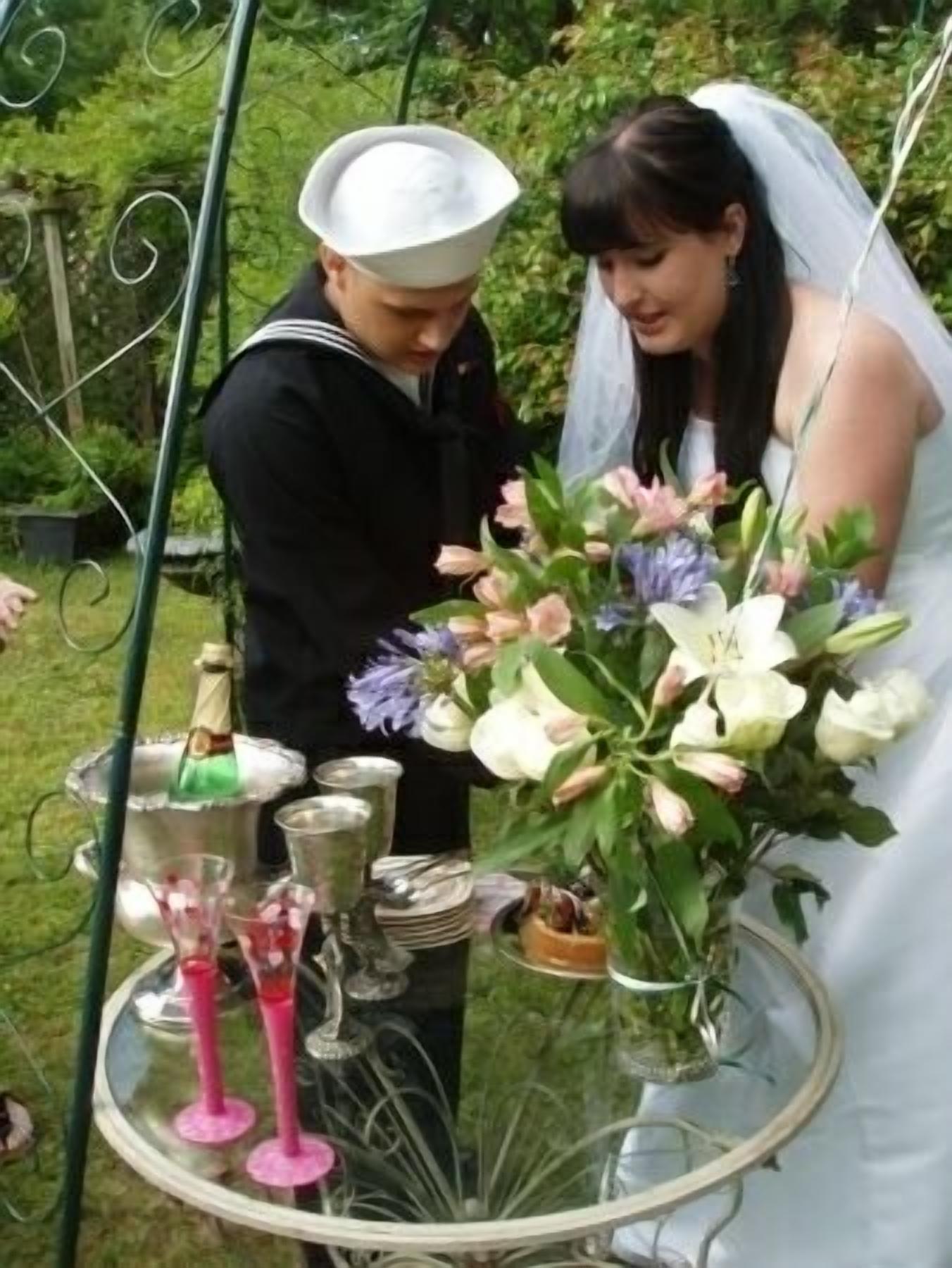 A wedding ceremony with a couple, a sailor, and a table with a champagne bottle, glasses, and flowers.
