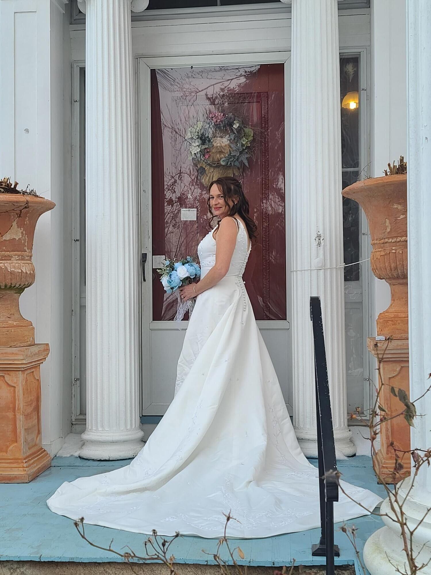 A bride standing in front of a door with a floral arrangement, columns, and a blue porch.