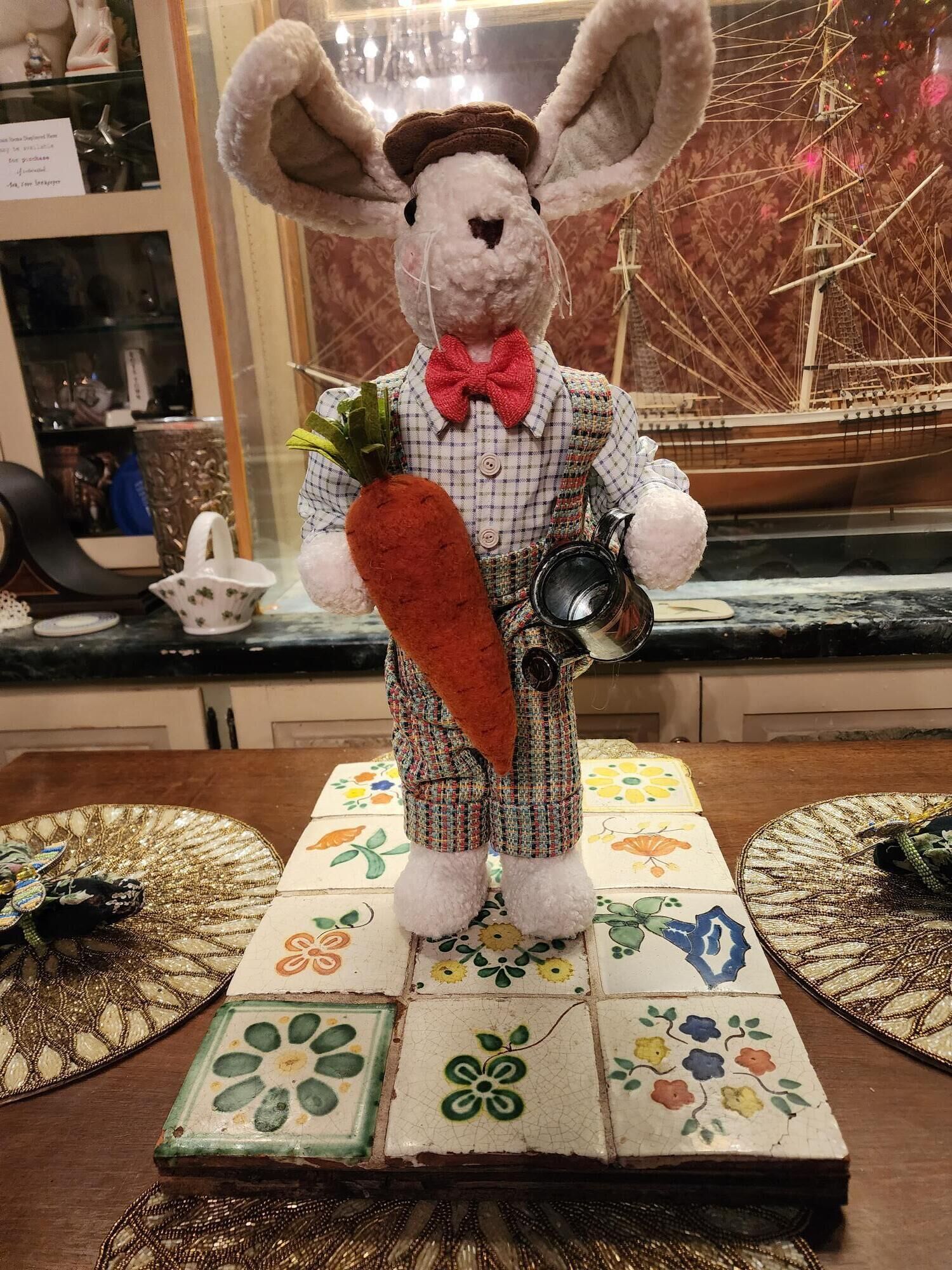 A plush rabbit figurine on a table with a patterned tablecloth, surrounded by various decorative items.
