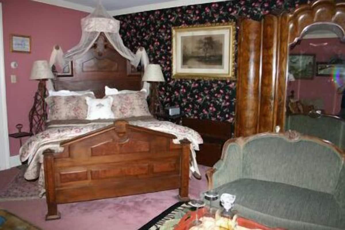 Ein Schlafzimmer mit einem hölzernen Himmelbett, einem grünen Sessel, floralem Tapetenmuster und einem gerahmten Bild an der Wand.