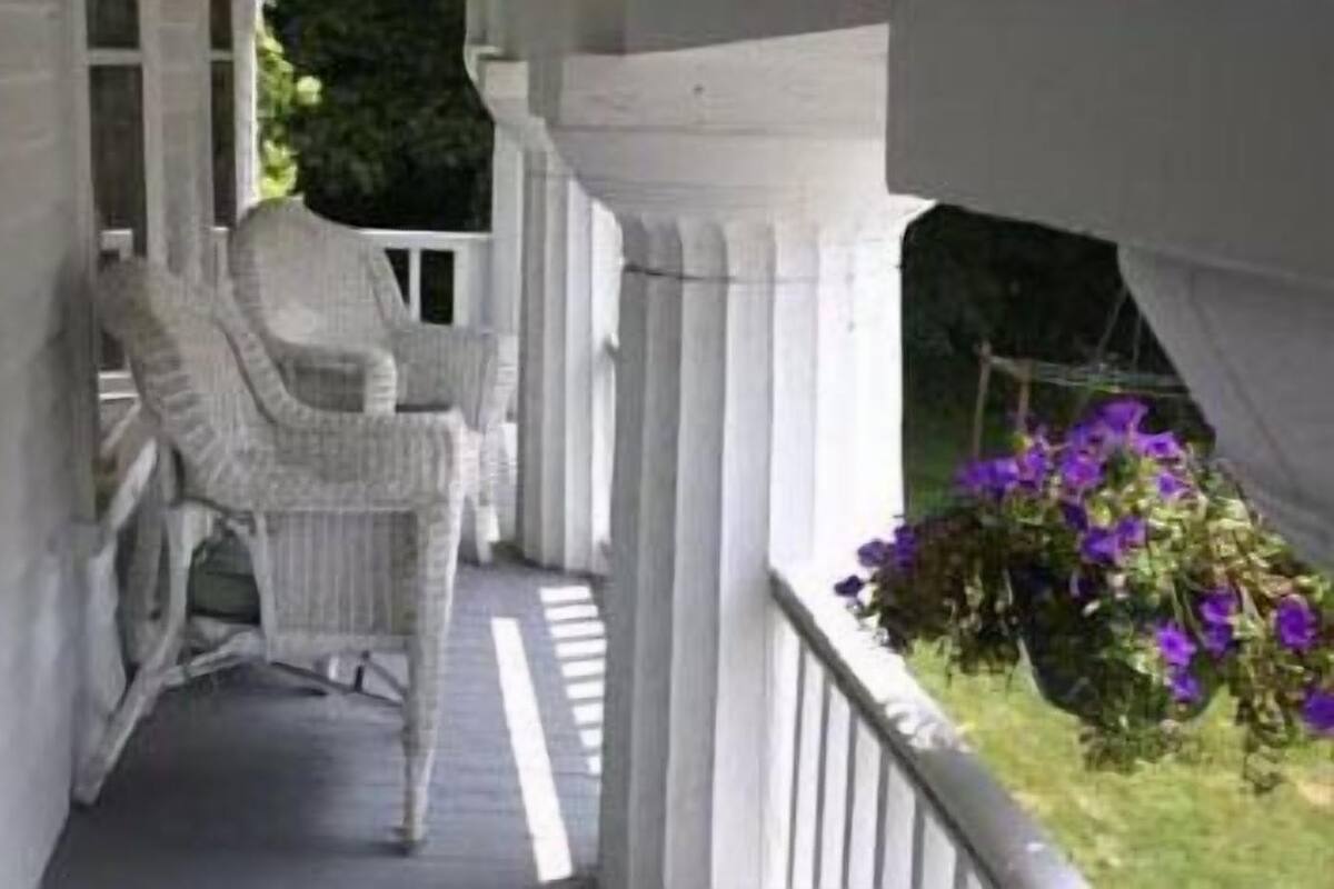 Eine Veranda mit zwei Korbstühlen, einem weißen Geländer und einem Blumenkasten mit violetten Blumen.
