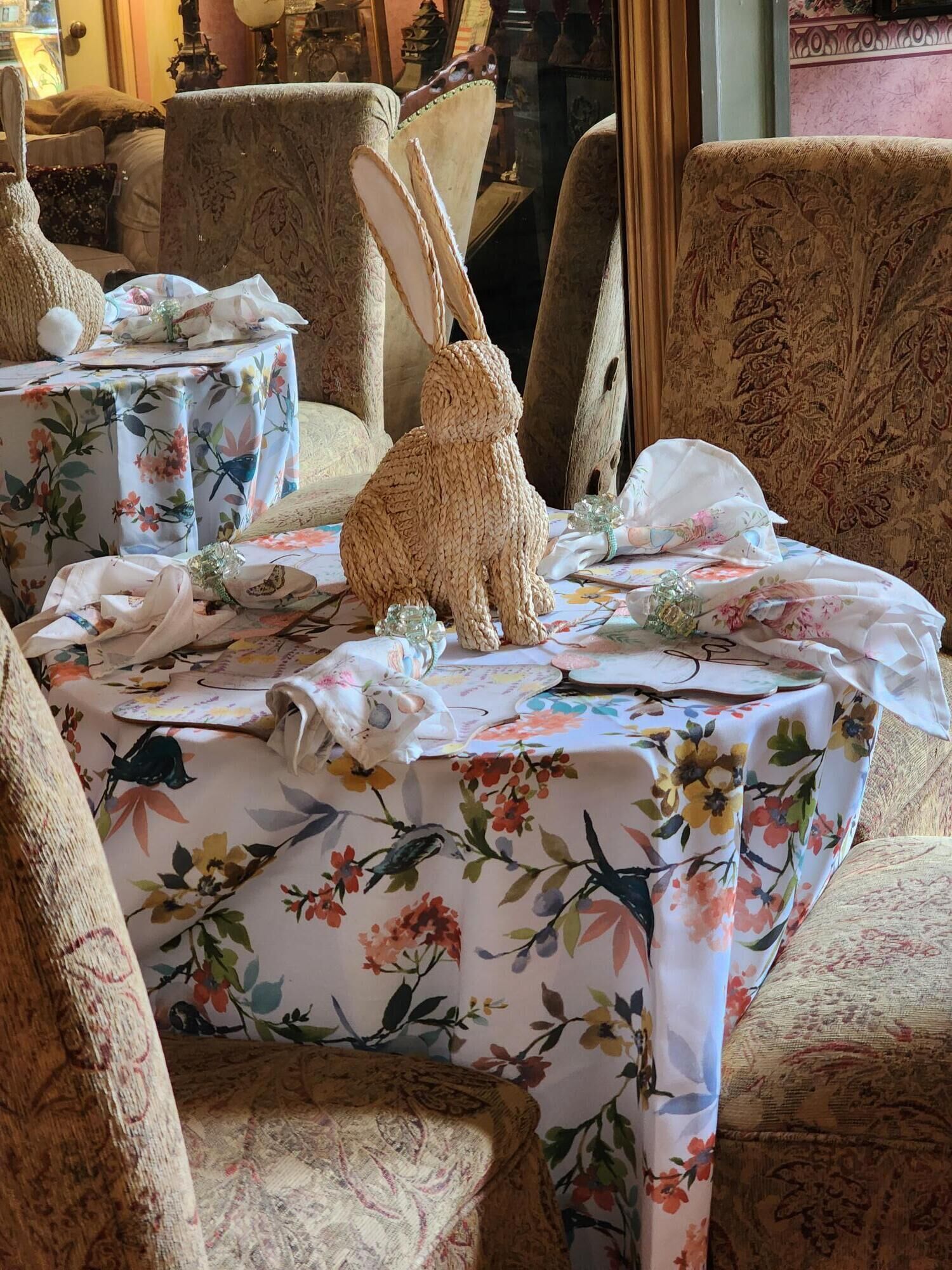 Una habitación con mesas y sillas de estampado floral, una decoración de conejo tejido y un espejo que refleja el interior de la habitación.