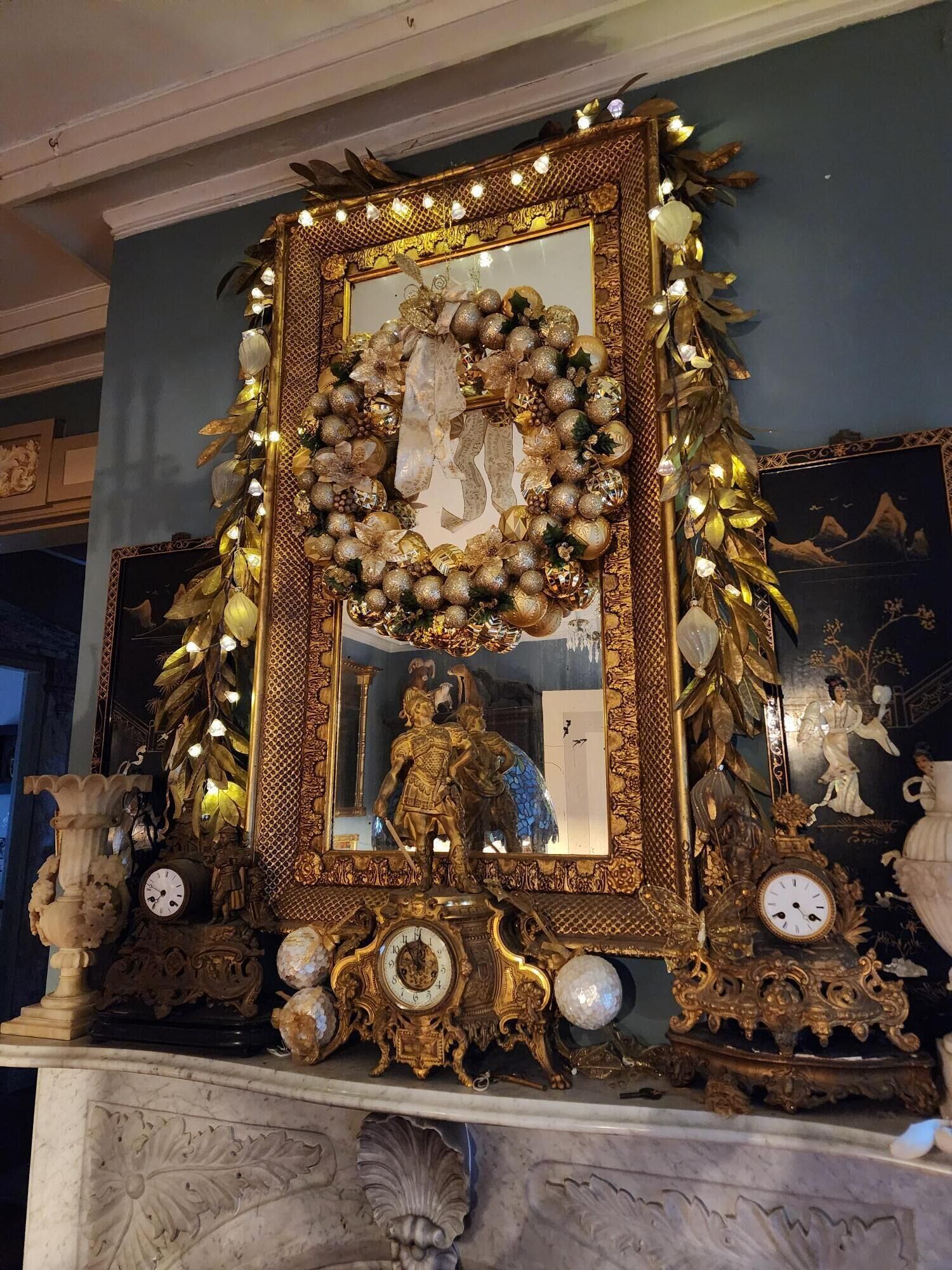 A grand mirror with a decorative wreath, surrounded by ornate gold and white decorations, set against a dark wall with classical artwork.