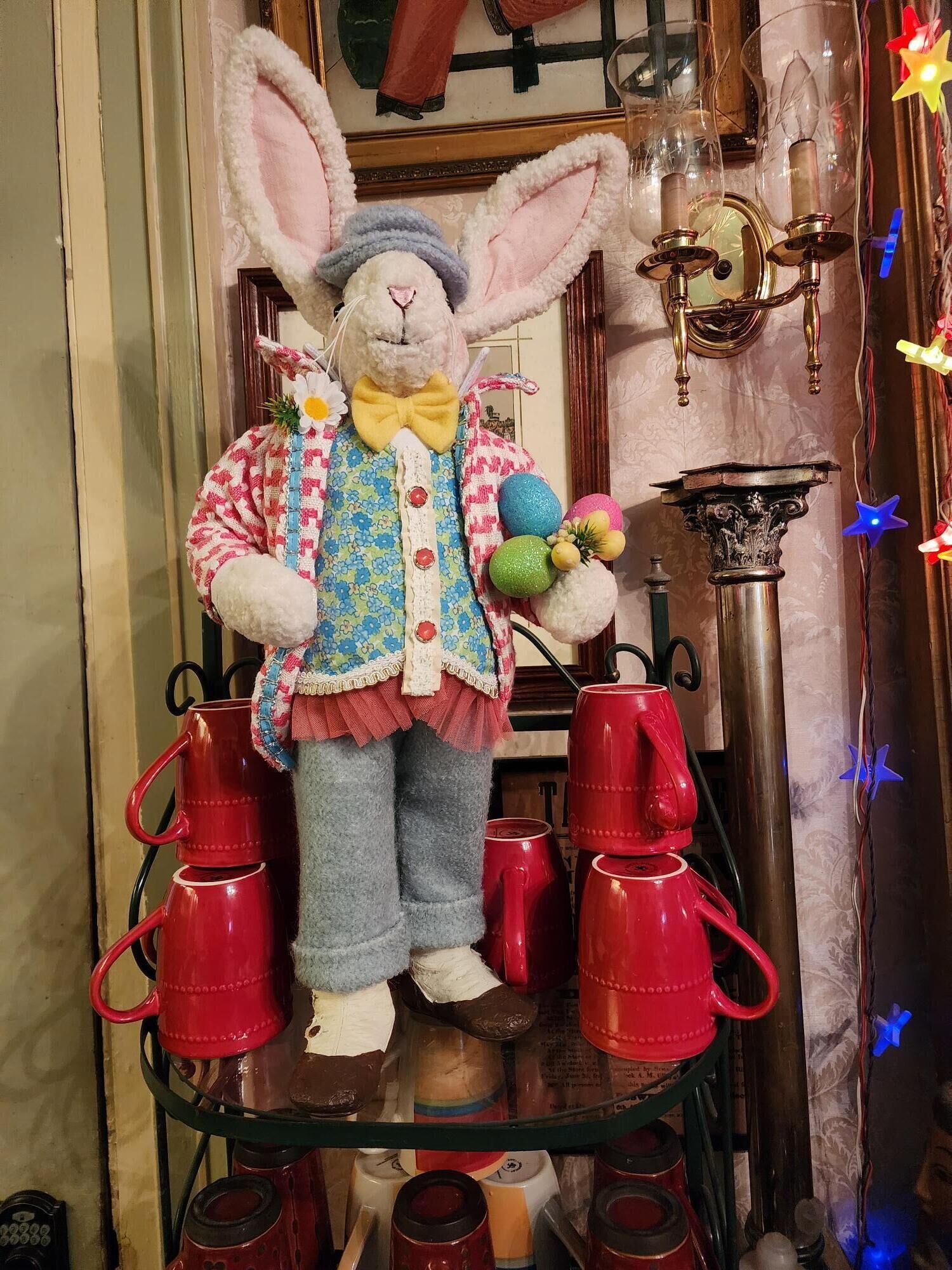 A plush rabbit figurine dressed in a patterned outfit, holding colorful eggs, and standing on a shelf with red mugs.