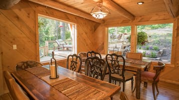 A wooden dining area with a long table and chairs, large windows, and a ceiling light fixture.